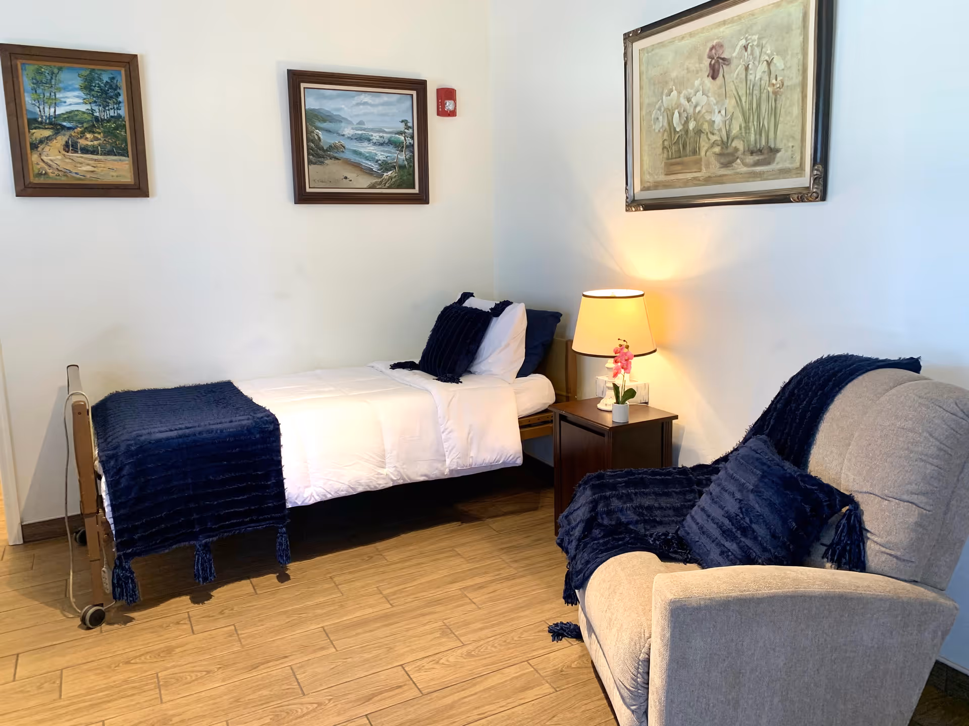 A small bedroom with a single bed covered in white bedding and a dark blue throw blanket and pillow. Next to the bed is a wooden nightstand with a lamp and a small pink flower in a pot. A beige armchair with a dark blue throw blanket and pillow is positioned nearby. The walls are decorated with three framed paintings, and the floor has a wood-like finish.