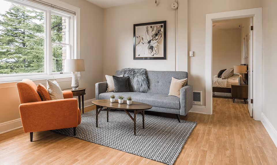 A cozy living room with a gray sofa adorned with pillows and a fur throw, an orange armchair with cushions, a wooden coffee table with three small potted plants, a side table with a lamp, a large window showing green trees outside, and a doorway leading to a bedroom with a bed and nightstand with a lamp.