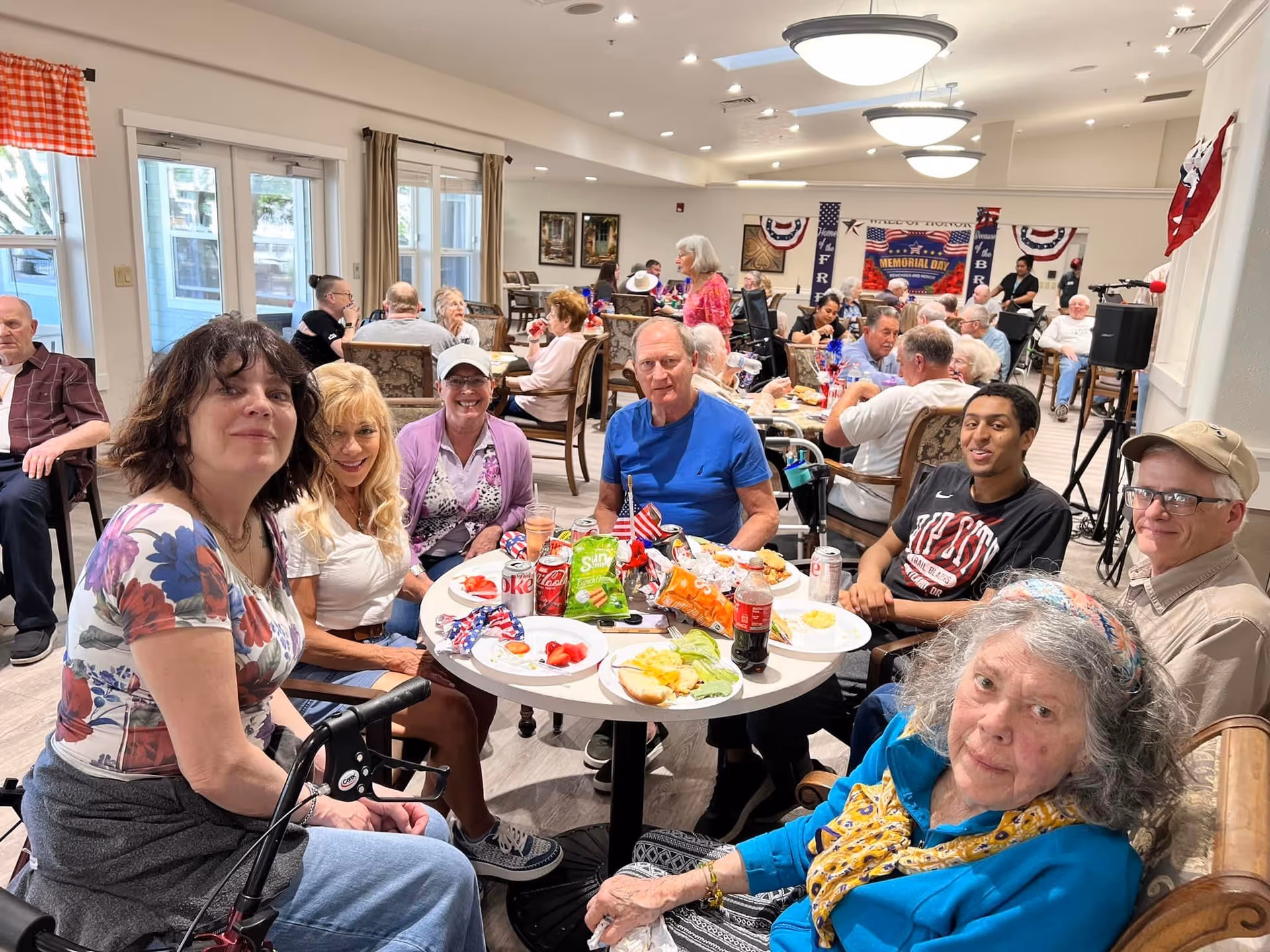 A group of elderly and middle-aged people sitting around a table in a well-lit dining area, enjoying a meal with plates of food, drinks, and snacks. The room is filled with other people seated at tables, and there are patriotic decorations on the walls, including banners and flags. The atmosphere appears social and lively.