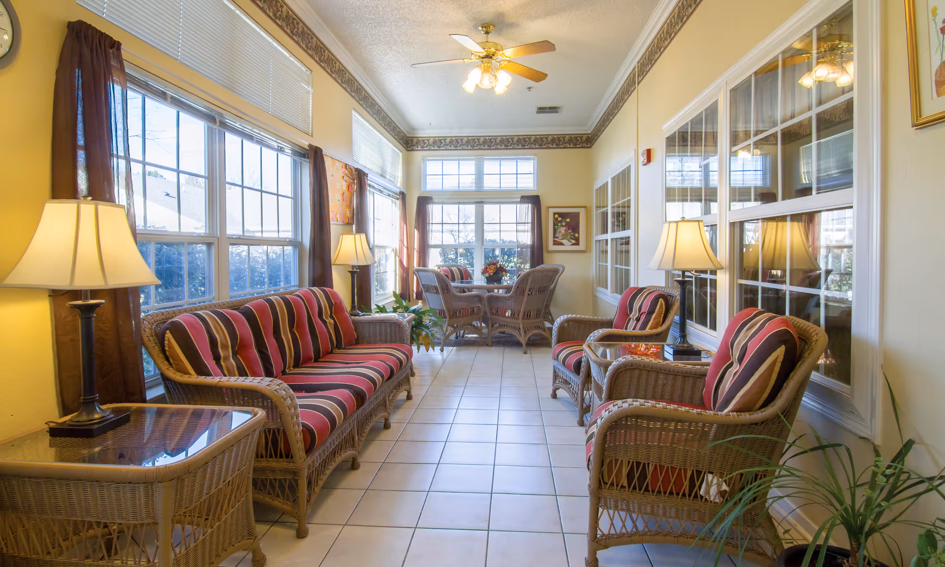 Sunlit common sitting area with wicker sofas and chairs with striped cushions, side tables and lamps along large windows.
