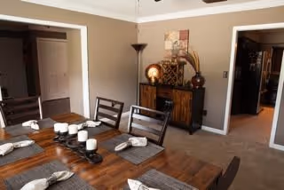 A dining room with a wooden table set for six people, each place setting having a napkin and a candle. The room has beige walls, a ceiling fan, a floor lamp, and a wooden sideboard with decorative items including a lamp and artwork above it. There are doorways leading to other rooms.