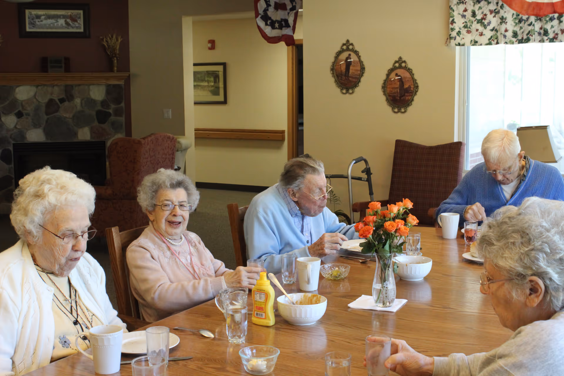 Five elderly individuals sitting around a wooden dining table in a senior living facility, enjoying a meal together. The table has glasses, cups, bowls, a bottle of mustard, and a vase with orange flowers. The room has a cozy atmosphere with a stone fireplace, armchairs, and wall decorations.