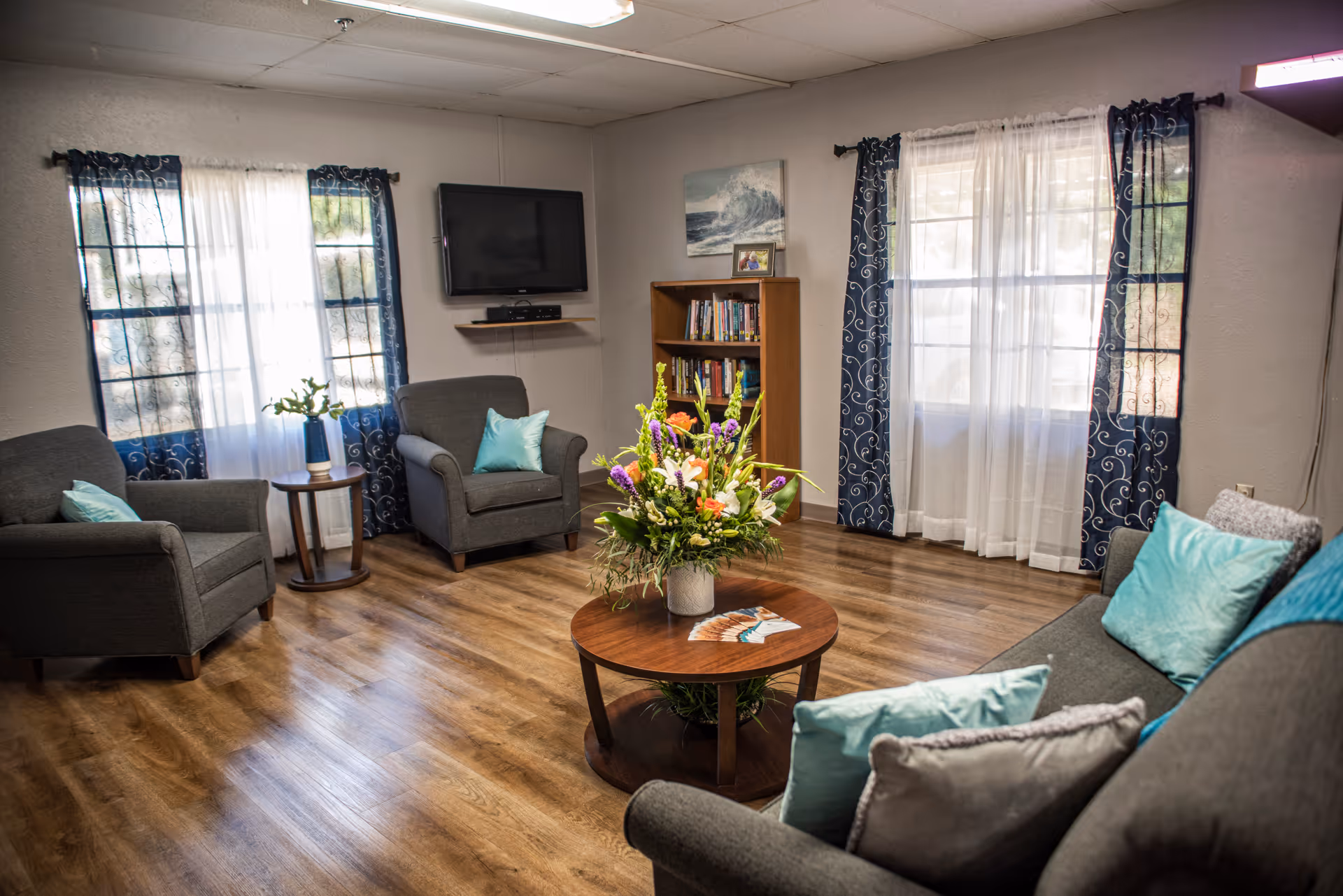 A cozy living room with wooden flooring, featuring a gray sofa and two gray armchairs with light blue and gray cushions. A round wooden coffee table in the center holds a colorful flower arrangement and some brochures. The room has two windows with sheer white curtains and dark blue patterned drapes. A small wooden side table with a vase of flowers is next to one armchair. A wall-mounted TV and a wooden bookshelf filled with books are visible on the far wall.