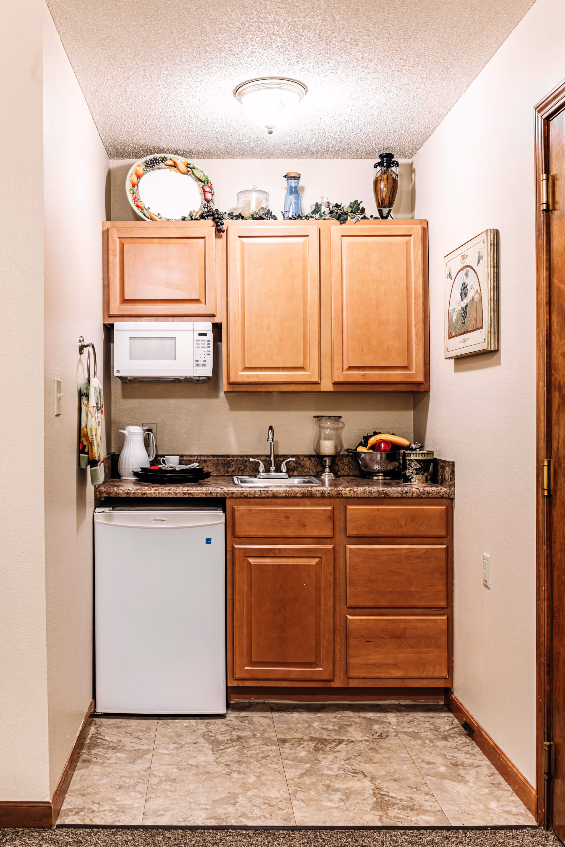 A small kitchenette with wooden cabinets, a white mini refrigerator, a microwave mounted under the upper cabinets, a sink with a faucet, and a countertop with a pitcher, plates, a bowl of fruit, and decorative items on top of the cabinets and on the wall.