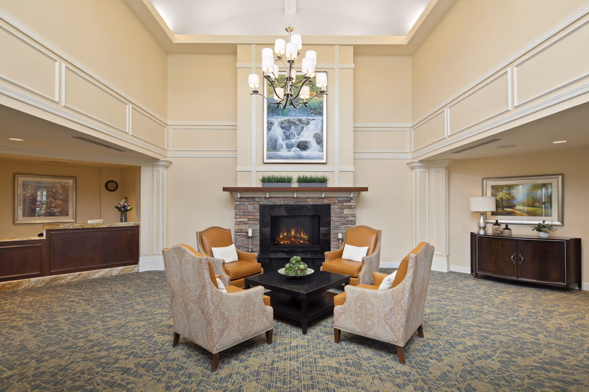 A cozy seating area in a senior living facility with four armchairs arranged around a square black coffee table. Behind the seating area is a stone fireplace with a lit fire, topped with a wooden mantel holding three small green plants. Above the fireplace hangs a framed picture of a waterfall. The room has beige walls with white trim, a high ceiling with a chandelier, and carpeted flooring. To the left is a reception desk with a vase of flowers, and to the right is a dark wooden cabinet with decorative items and a framed landscape painting above it.