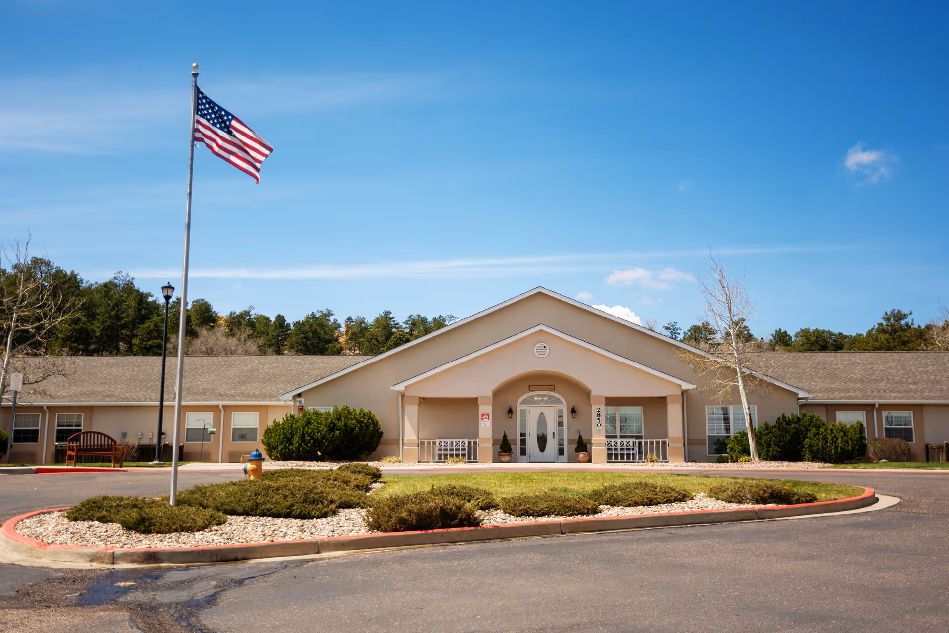 Front exterior view of a single-story senior living facility building with a beige facade, a peaked roof, and a covered entrance. There is an American flag on a flagpole in front of the building, surrounded by landscaped bushes and a circular driveway. The sky is clear and blue.