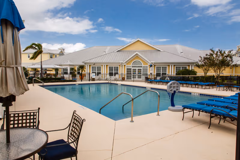 Outdoor swimming pool area with blue lounge chairs and patio furniture in front of a yellow senior living building.