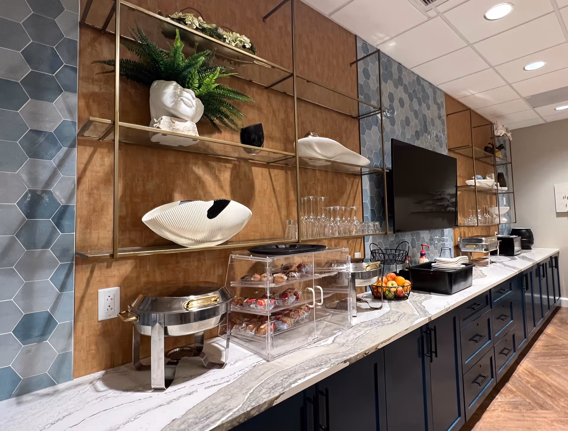 A modern buffet counter with a marble countertop featuring a variety of items including a chafing dish, a clear display case with pastries, a fruit basket, glasses, and napkins. The wall behind has decorative shelving with plants and art pieces, a mounted flat-screen TV, and hexagonal tile accents.