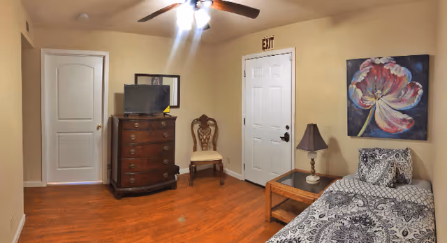 A cozy room with wooden flooring, a single bed with patterned bedding, a wooden side table with a lamp, a floral painting on the wall, a wooden chair, a chest of drawers with a TV on top, and a ceiling fan with lights. There is a white door with an exit sign above it and another closed door on the left side.