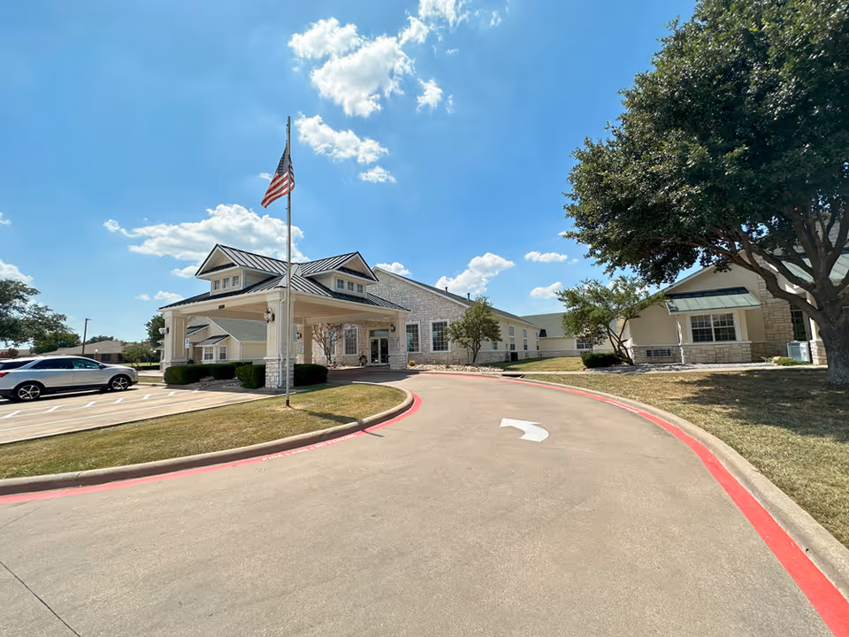 Exterior view of Colonial Lodge Assisted Living facility on a sunny day, showing the driveway with a curved red curb, an American flag on a flagpole, parked cars, and the building surrounded by trees and landscaping.