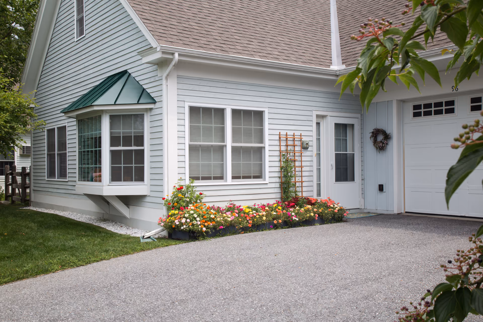 Exterior view of a light blue house with white trim, featuring a bay window with a green metal awning, a flower bed with colorful flowers along the wall, a white door, and a garage door with a wreath. The driveway and green lawn are visible in the foreground.