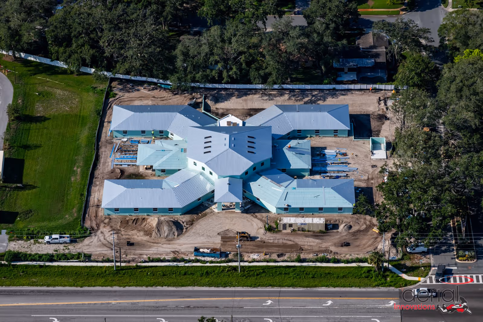 Aerial view of a large building under construction surrounded by dirt and some greenery, with a road in the foreground and trees in the background.