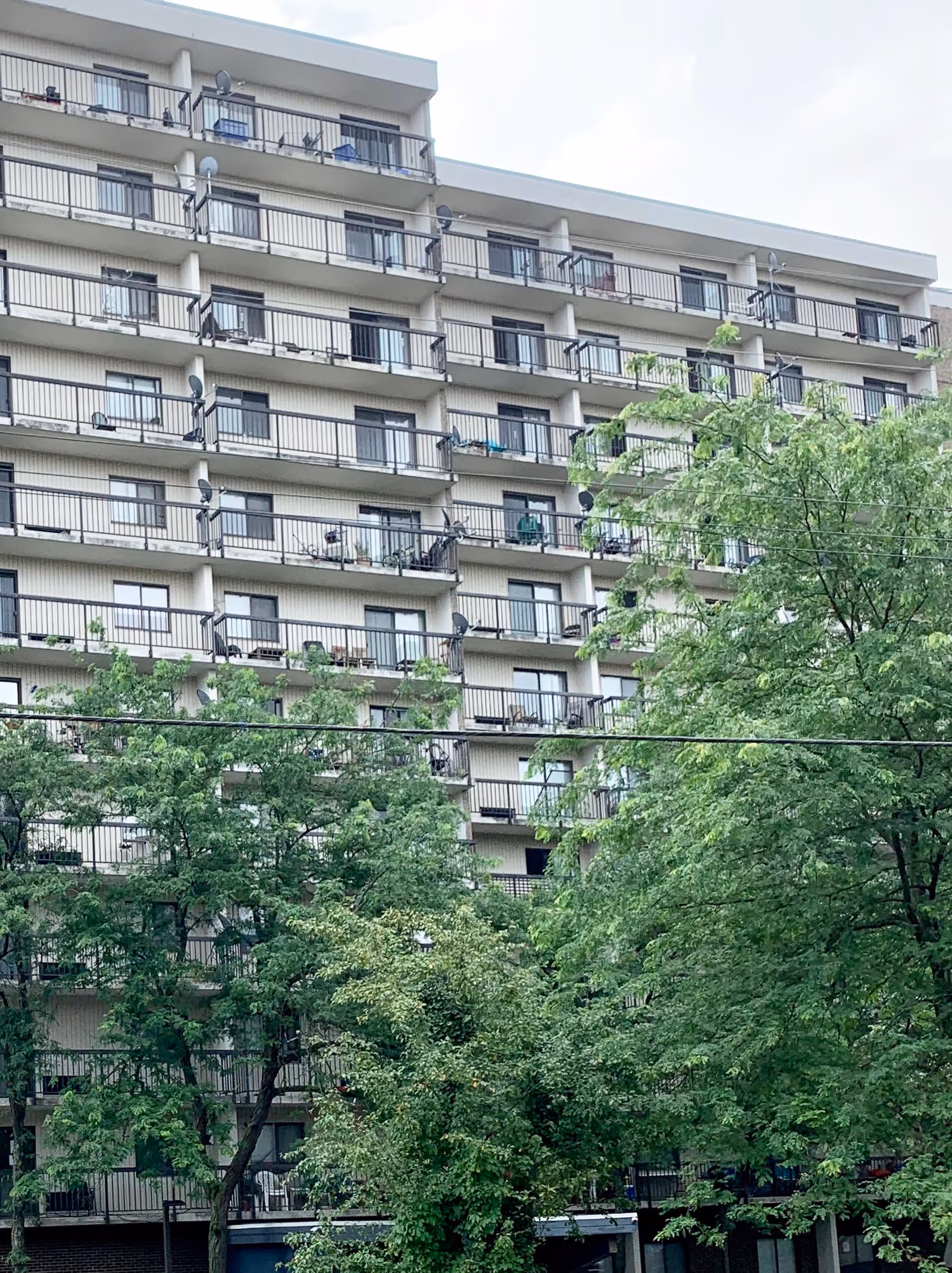 Exterior view of a multi-story senior living facility building with balconies on each floor. The building is partially obscured by green trees in the foreground under a cloudy sky.