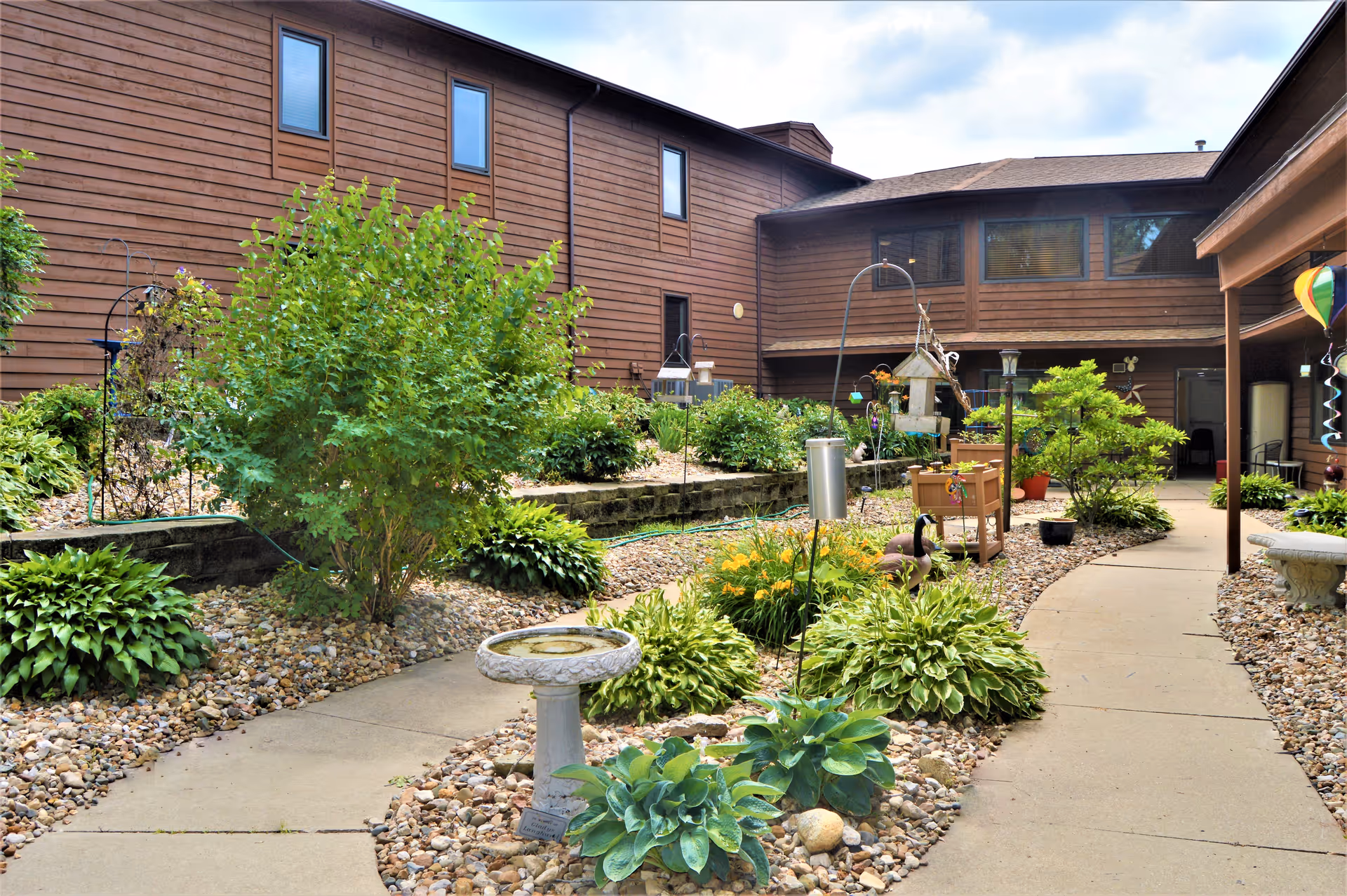 A landscaped courtyard garden area with a concrete pathway winding through various green plants, shrubs, and flowers. The courtyard is surrounded by a two-story brown wooden building with several windows. There are decorative elements such as a birdbath, bird feeders, and garden ornaments along the pathway.