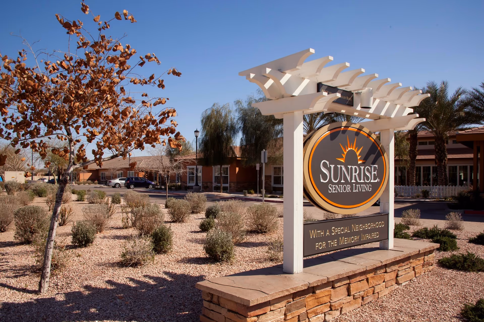 Outdoor view of Sunrise Senior Living facility in Chandler, featuring a large sign with the facility's name and a message about a special neighborhood for the memory impaired. The area around the sign has desert landscaping with small bushes and a tree with brown leaves, under a clear blue sky.