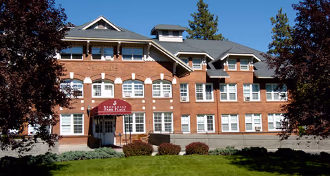 Front exterior view of a large brick building with multiple windows and a maroon awning above the entrance that reads 'Brookdale Park Place'. The building is surrounded by green grass and trees with a clear blue sky above.
