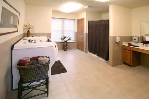 A spacious bathroom with a large white bathtub on the left side, a wicker basket with rolled towels in front of it, a brown bath mat on the tiled floor, a window with blinds, a door, a shower area with a dark curtain, and a vanity with a sink and mirror on the right side. The walls are light-colored with beige tiles halfway up.