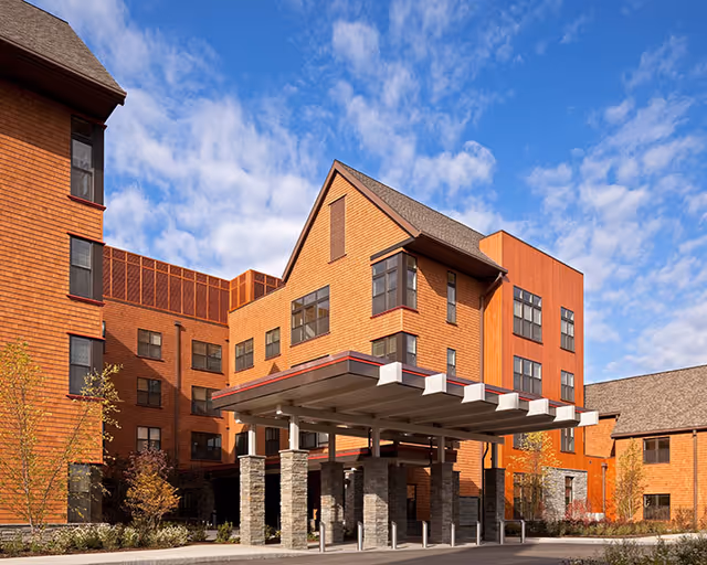 Exterior view of a multi-story assisted living facility building with a covered entrance supported by stone pillars. The building features a combination of wood and brick siding under a partly cloudy blue sky.