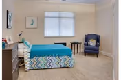 Bright, simply furnished bedroom with a single bed, dresser, armchair, and a window with blinds.