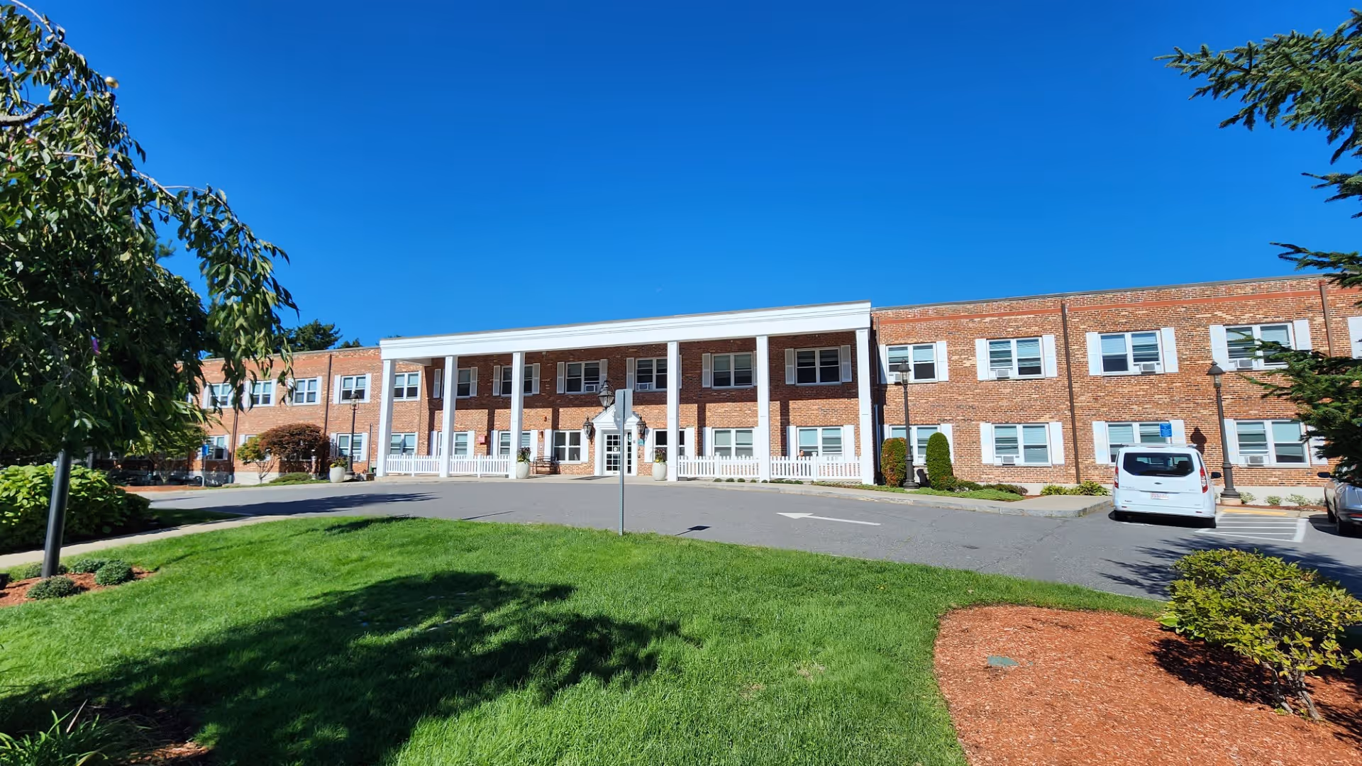 Two-story red-brick senior living building with white columns at the main entrance, front lawn, and parked cars under a clear blue sky.