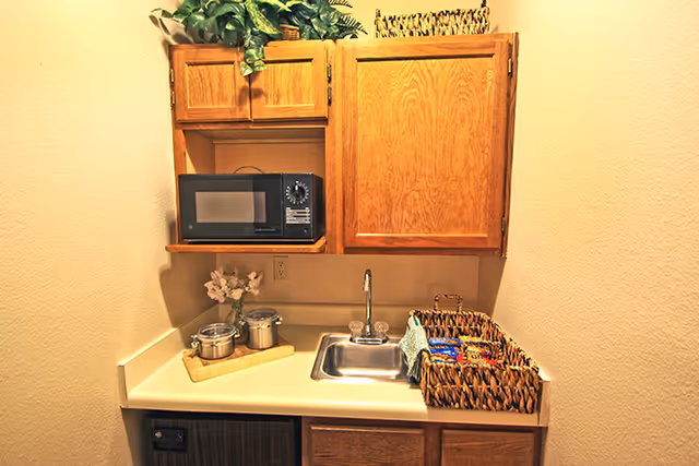 Small kitchenette area with wooden cabinets, a microwave, a stainless steel sink, a basket filled with snacks, and a small tray with containers and a flower vase on the countertop.