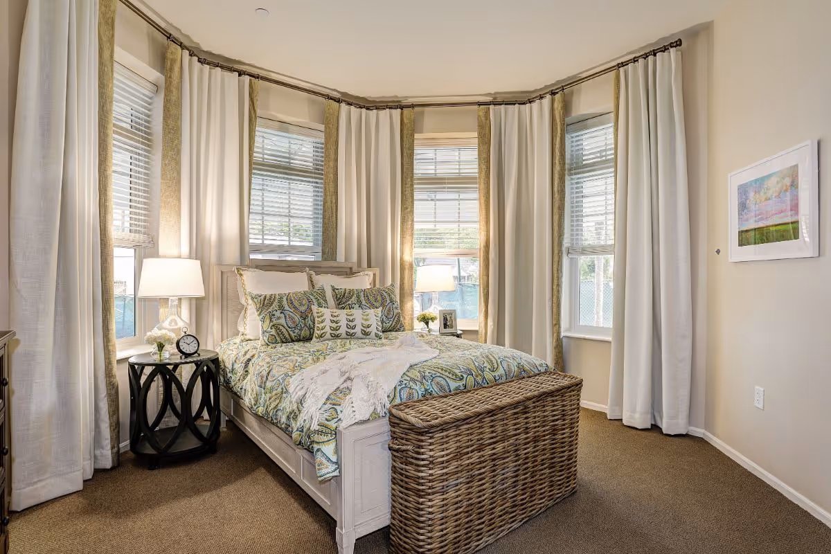 A bright bedroom with a bed centered in a bay of windows, patterned bedding, bedside tables with lamps, and a woven storage bench at the foot of the bed.