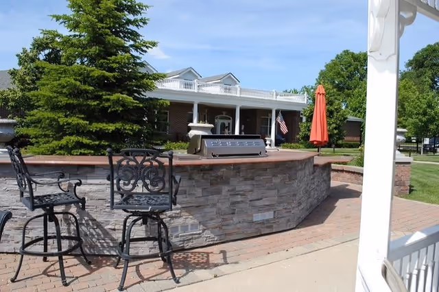 Outdoor patio area with a stone bar counter, two ornate black metal bar stools, a grill on the counter, a red umbrella, and a large evergreen tree. In the background, there is a brick building with white columns and an American flag.