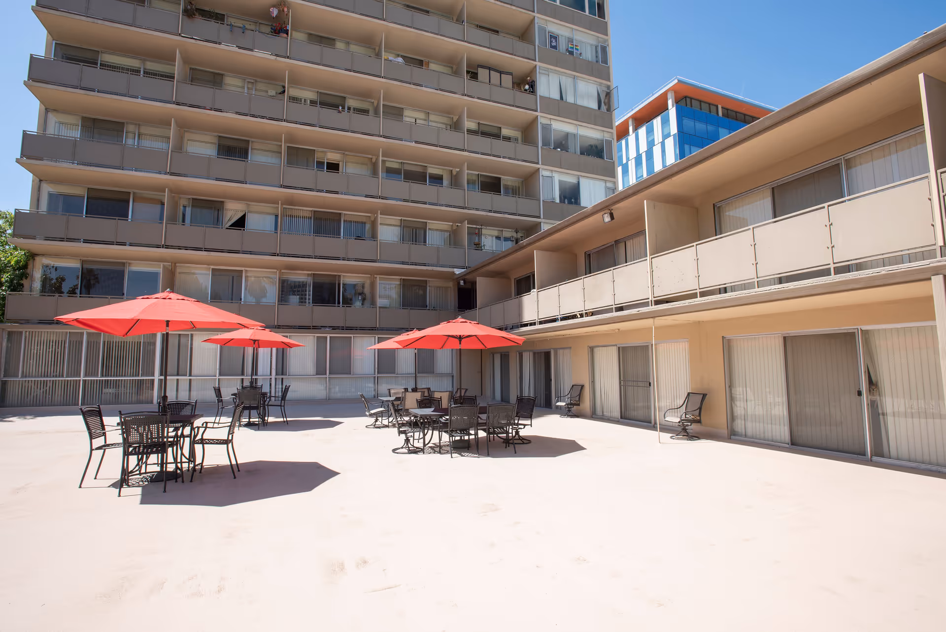 Outdoor patio area at Park Place Apartments with several black metal tables and chairs, each shaded by red umbrellas. The patio is surrounded by a multi-story building with balconies and sliding glass doors. A modern blue and orange building is visible in the background under a clear blue sky.