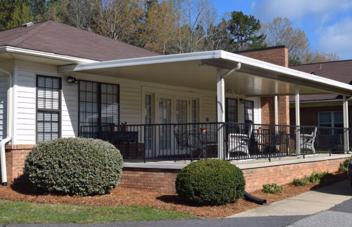 Covered front porch with black metal railing and patio chairs on a single-story brick-and-siding building with shrubs in front.