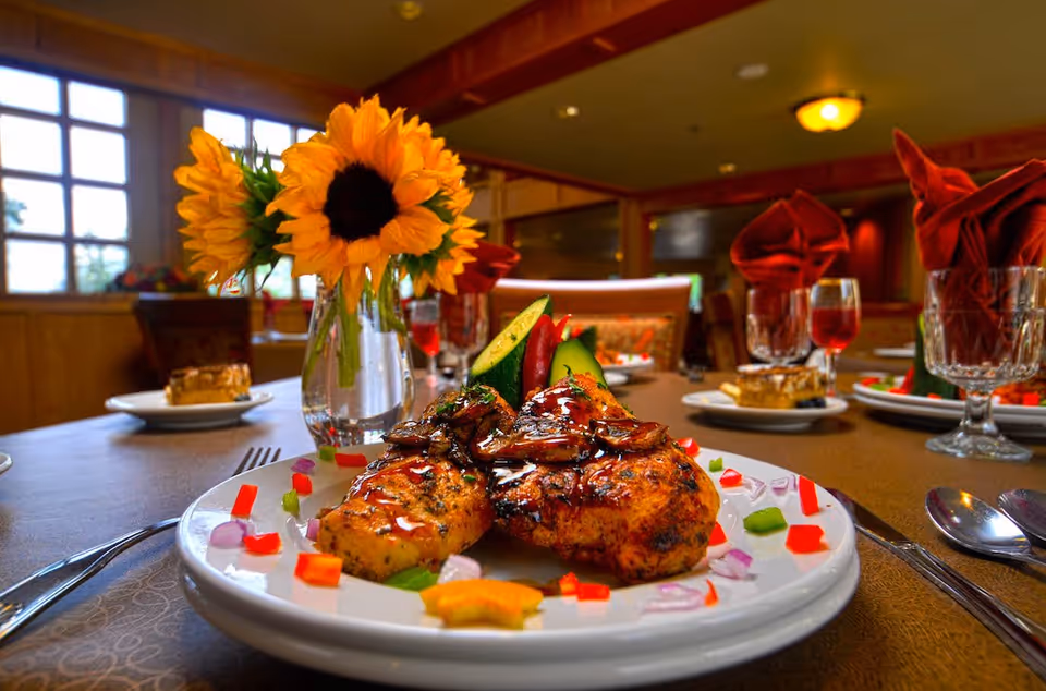 Plated roasted chicken with garnish on a dining table set with glasses, napkins, and a vase of sunflowers in a dining room.