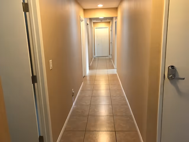 Long tiled interior hallway with beige walls and multiple doors on each side.