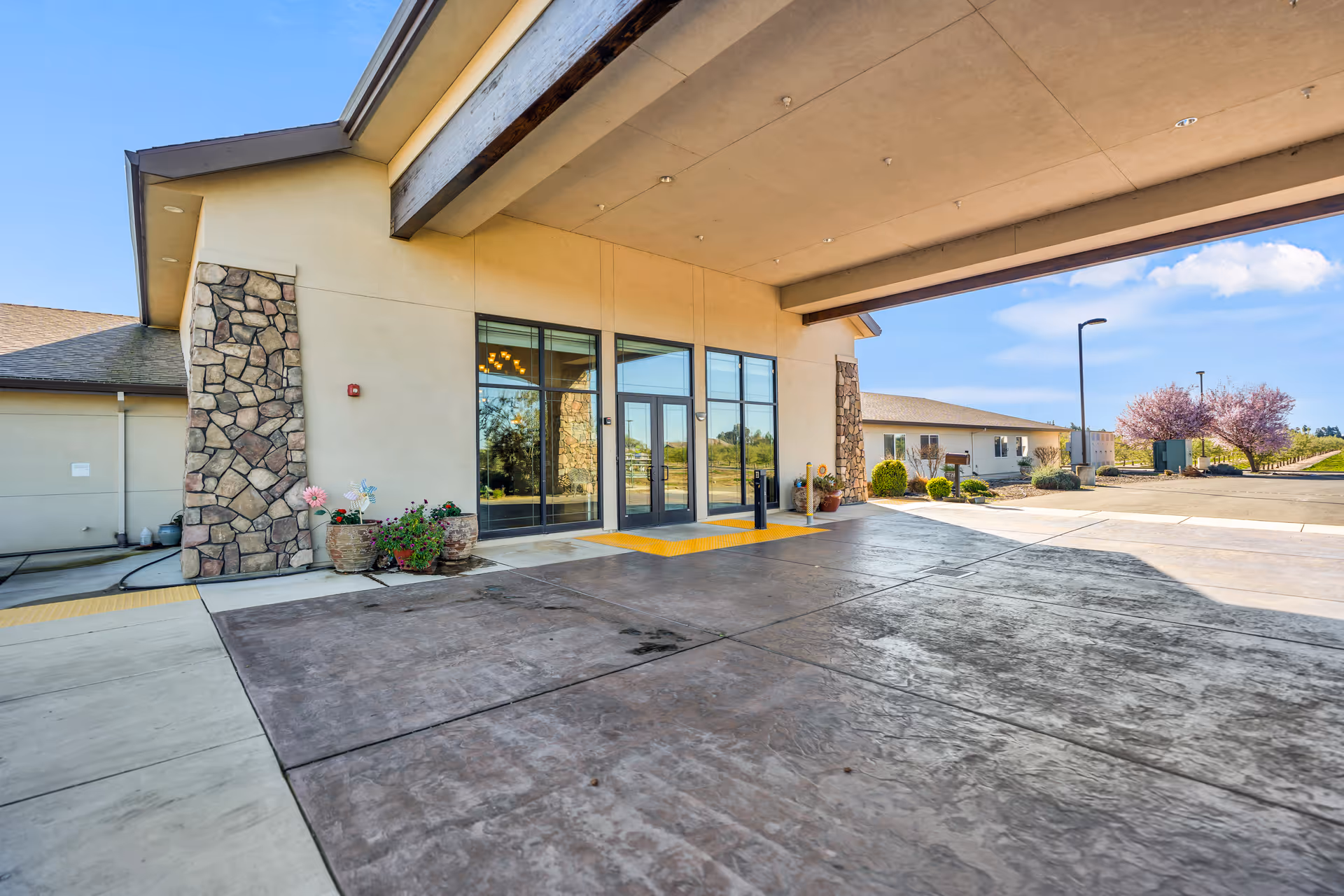 Covered entrance and drop-off area of a senior living facility with stone pillars, glass double doors, potted plants, and a driveway leading to landscaped grounds.