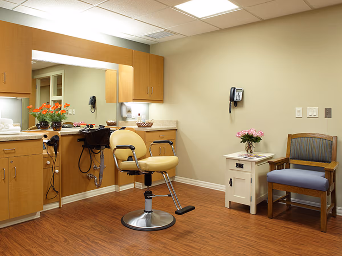 A well-lit room with wooden cabinets and a large mirror above the counter. There is a yellow salon chair in front of the counter, a hair dryer attached to the cabinet, and a black sink for washing hair. On the right side, there is a small white side table with a vase of pink flowers and a wooden chair with a blue cushion. The room has wood flooring and beige walls with a wall-mounted telephone.