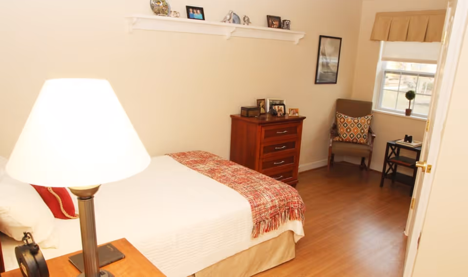 A cozy bedroom with a neatly made bed covered with a white bedspread and a red patterned throw blanket. A wooden nightstand with a lamp and an alarm clock is beside the bed. Across from the bed is a wooden dresser with framed photos on top. A cushioned chair with a decorative pillow sits near a window with a beige valance and a small side table with a plant and a book. The room has wooden flooring and light-colored walls with a small shelf displaying decorative items.