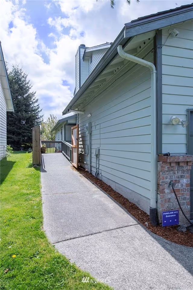 Outdoor view of a light blue building with a concrete pathway leading to a wooden gate. The pathway is bordered by green grass on one side and the building on the other. A small sign near the building reads 'PARKLANE AFH Entrance'. The sky is partly cloudy.