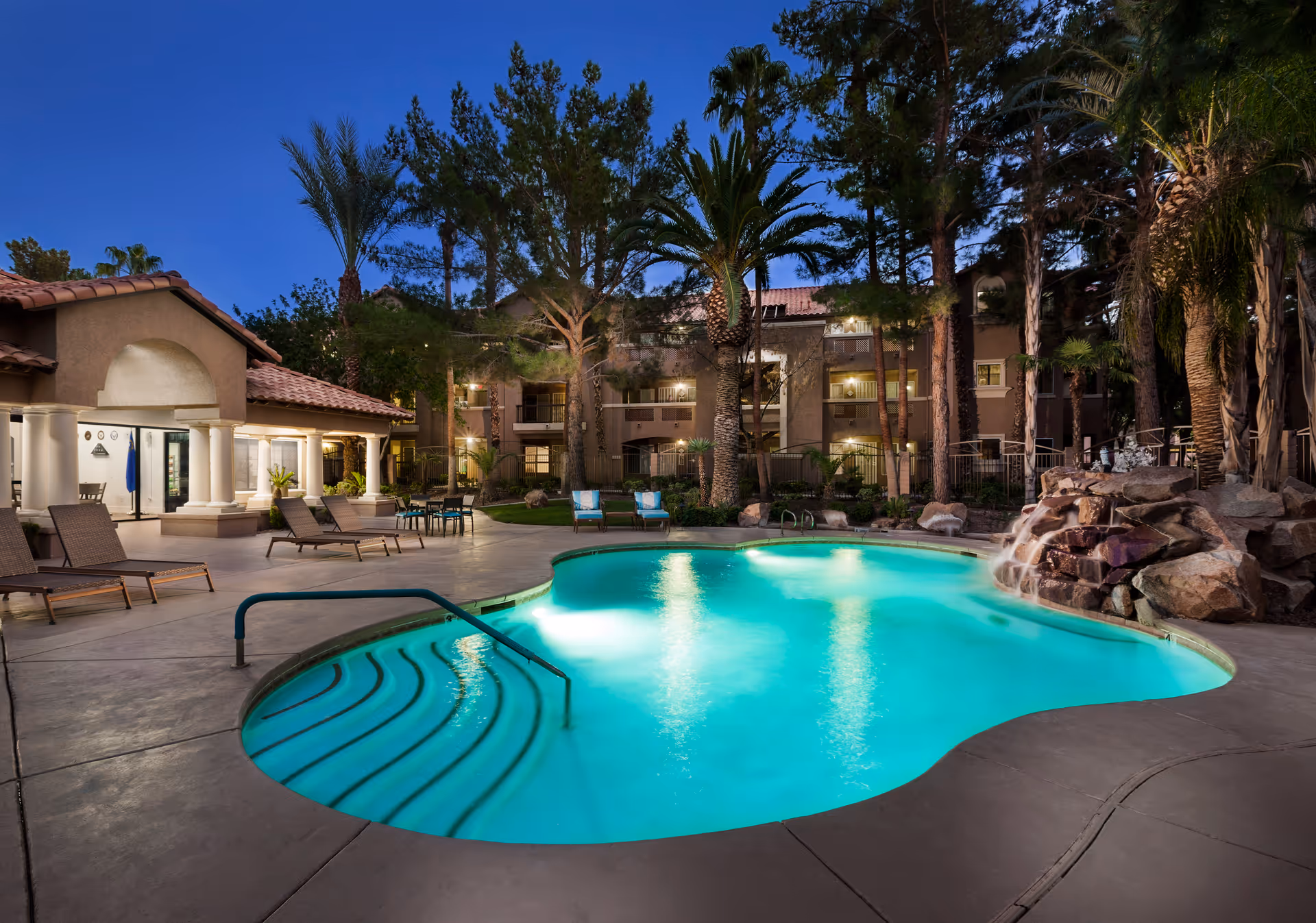 Outdoor swimming pool area at dusk with illuminated water, surrounded by lounge chairs, tables, palm trees, and a building with balconies and warm lighting in the background.