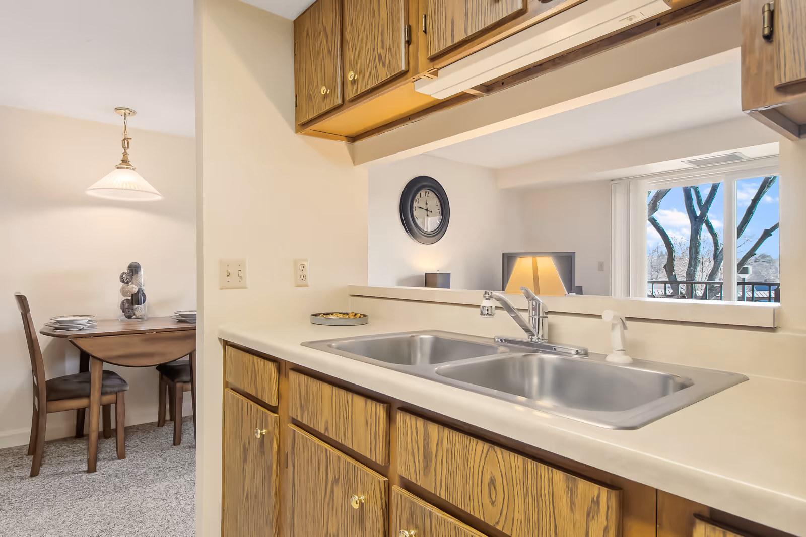 Interior view of a kitchen area with a double stainless steel sink and wooden cabinets. In the background, there is a small dining area with a wooden table set for four and a hanging light fixture. A wall clock and a lamp are visible in the adjacent living space, along with a window showing tree branches outside.