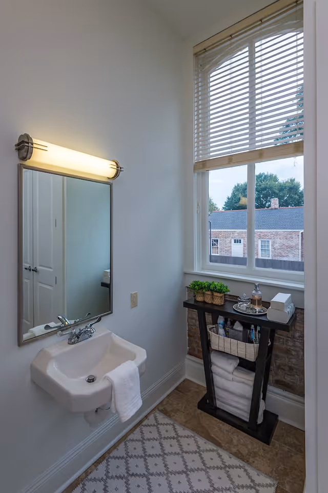 A small bathroom with a wall-mounted sink and a white towel hanging on it. Above the sink is a rectangular mirror with a horizontal light fixture. To the right, there is a window with blinds partially open, showing a view of a brick building and trees outside. Below the window is a small black shelf holding a potted plant, toiletries, and neatly folded white towels. The floor has a patterned rug.