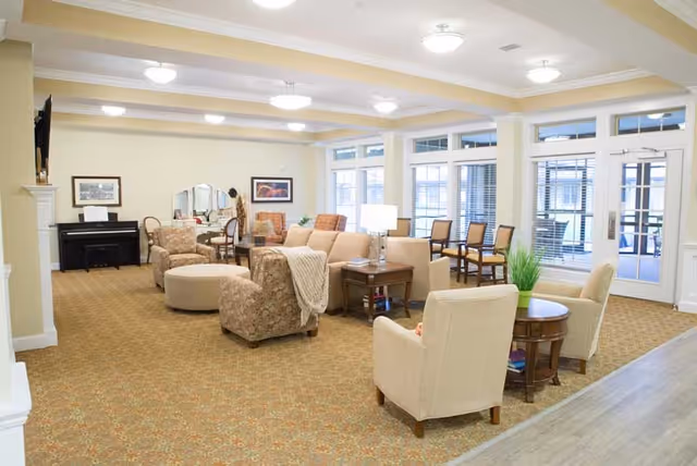 A spacious and well-lit living room area in a senior living facility with multiple armchairs, a sofa, side tables with lamps, a round ottoman, a piano, and large windows and glass doors letting in natural light.