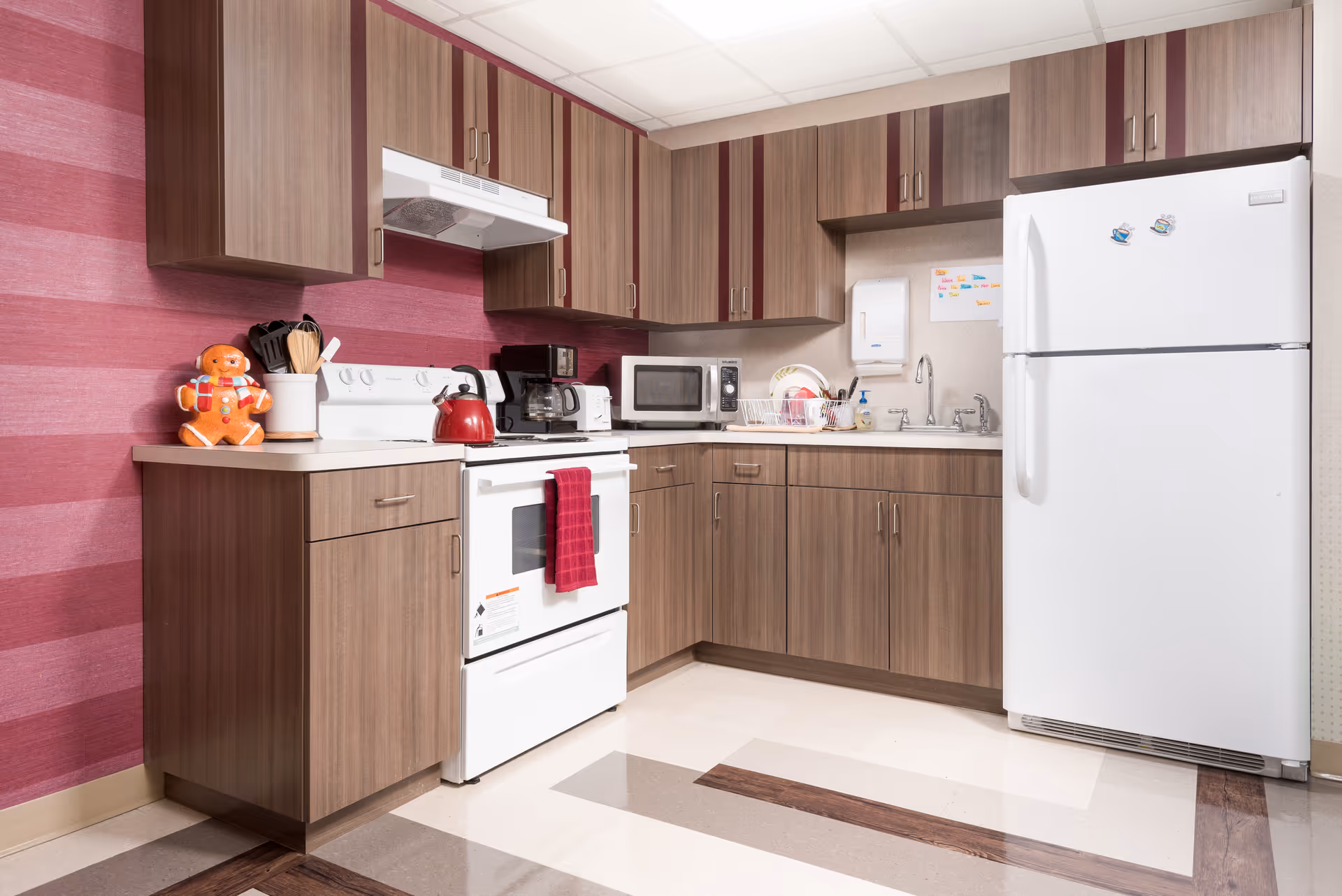 A compact kitchen with wood-tone cabinets, a white stove and refrigerator, microwave, sink, and pink striped wallpaper.