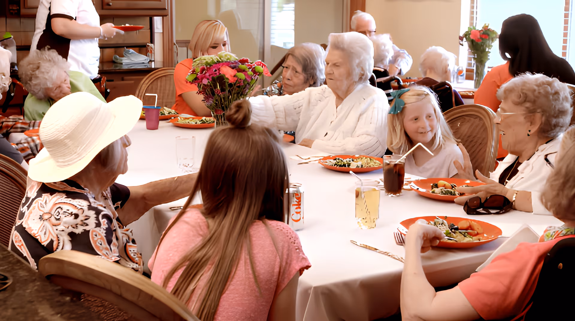 A group of elderly people and a few younger individuals sitting around a large dining table in a well-lit room, enjoying a meal together. The table is covered with a white tablecloth and has plates of food, drinks, and a vase with colorful flowers in the center. A staff member is serving food in the background.