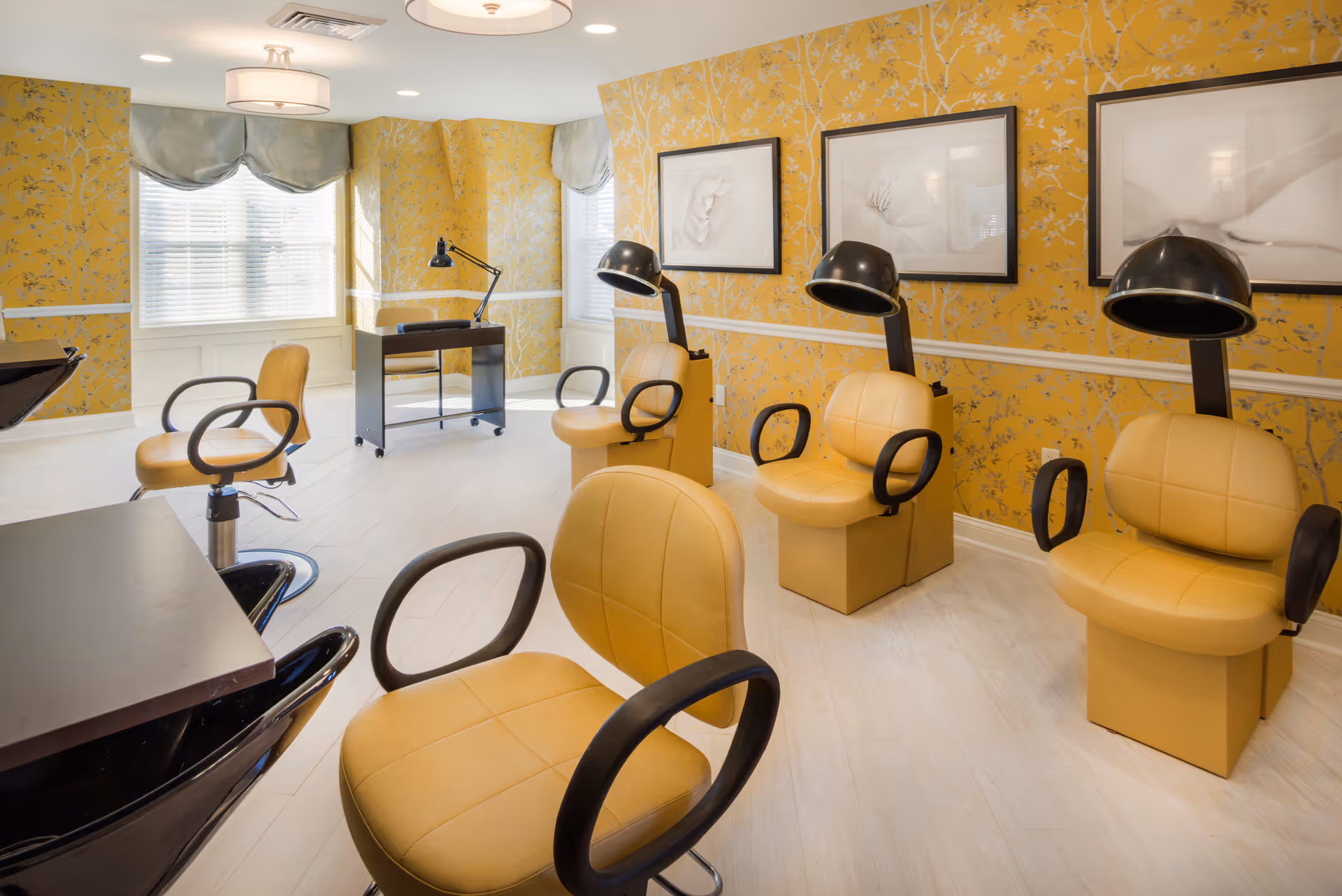 A bright salon area with mustard yellow salon chairs and hair drying stations against a yellow floral wallpapered wall. The room has large windows with gray valances, light wood flooring, and framed artwork on the walls. There is also a small black desk with a lamp in the background.