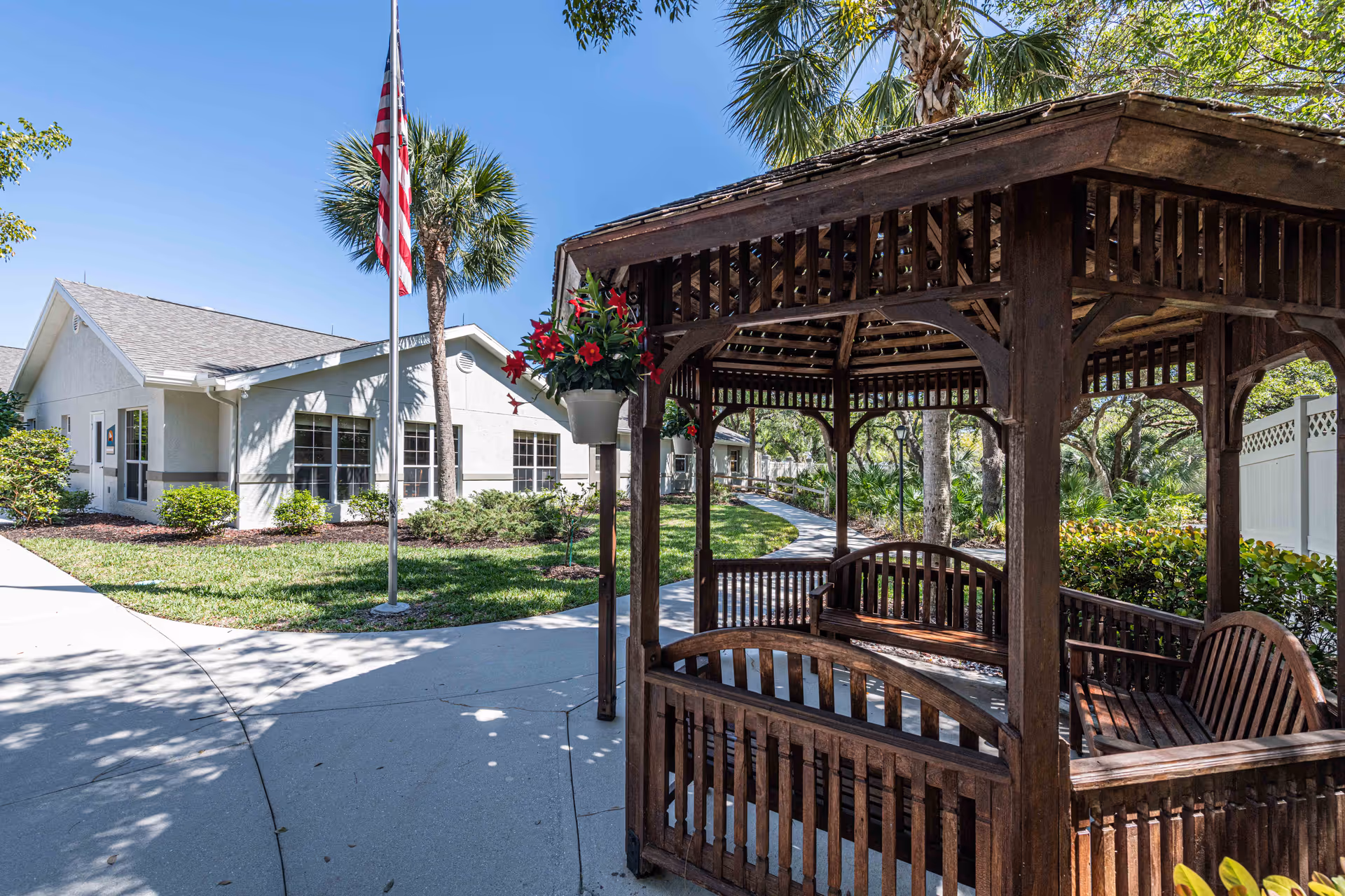 Outdoor area of Arden Courts - ProMedica Memory Care Community in Ft. Myers featuring a wooden gazebo with benches, a concrete pathway, an American flag on a flagpole, palm trees, and a white building in the background under a clear blue sky.