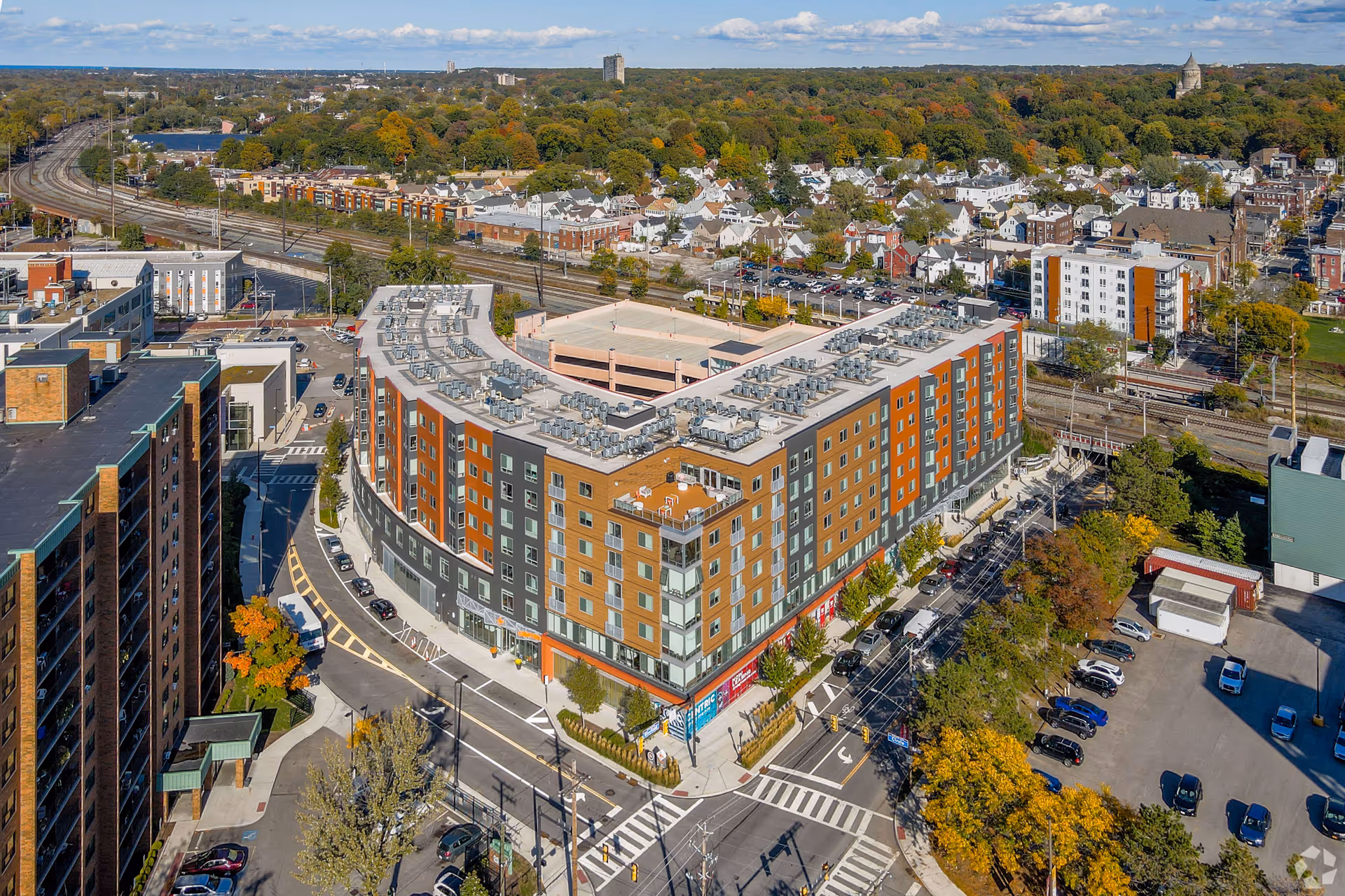 Aerial view of a large multi-story building with a U-shaped design, surrounded by streets, parked cars, and trees with autumn foliage. The building has a flat roof with numerous HVAC units and features a mix of brown, orange, and gray exterior panels. Residential houses and other buildings are visible in the background under a partly cloudy sky.