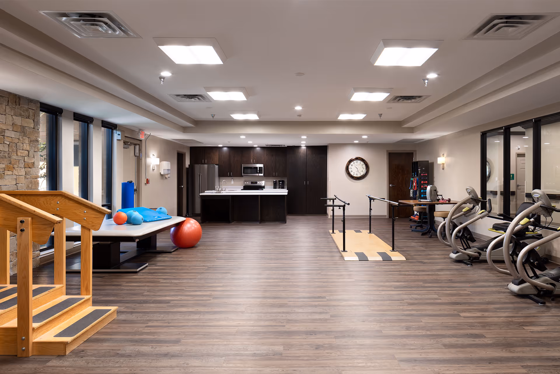 A spacious rehabilitation or physical therapy room with wooden stairs for practice, exercise balls, parallel bars for walking support, and several exercise machines. The room has a modern kitchen area with dark cabinets, a refrigerator, microwave, and sink at the back. The floor is wood-style, and the room is well-lit with ceiling lights and large windows on the left side.