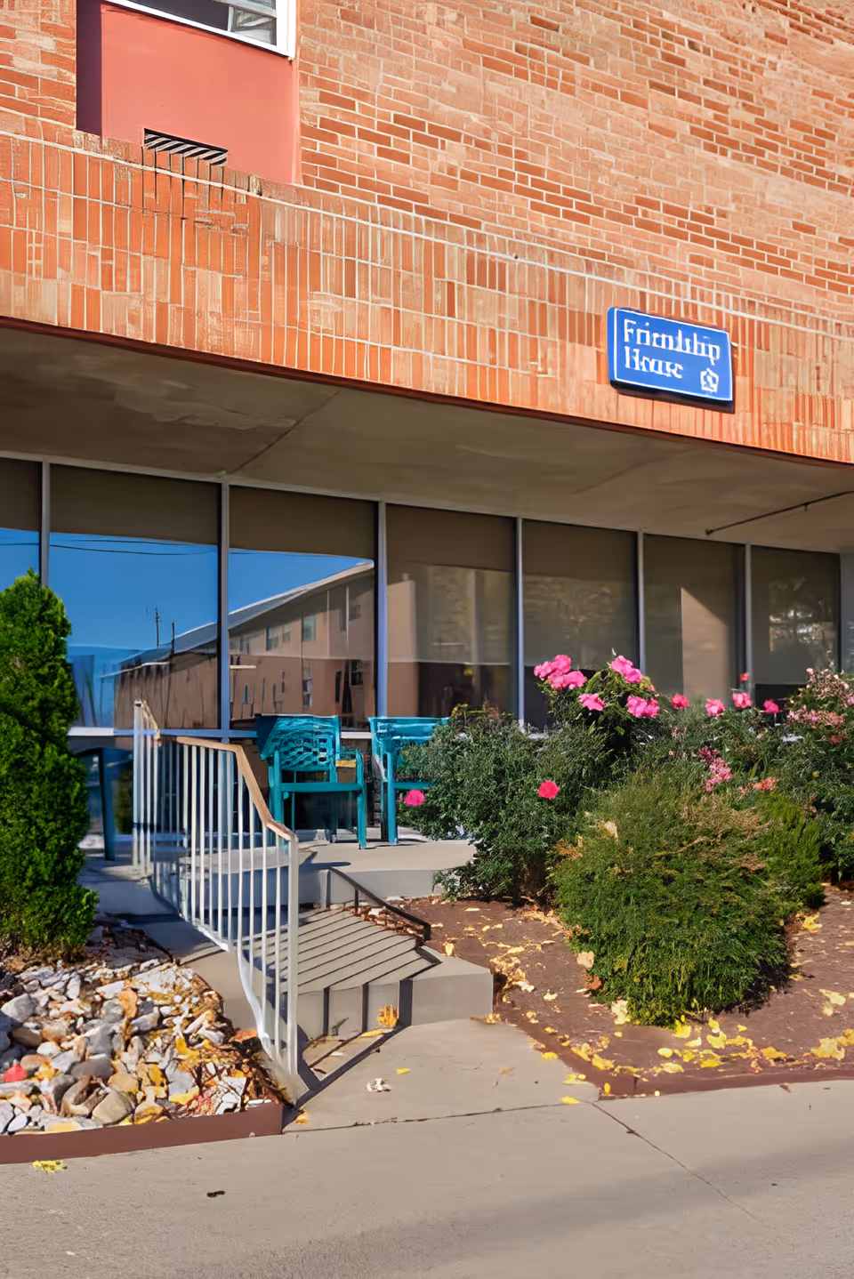 Outdoor entrance area of a building with a small staircase and ramp leading to a patio with teal chairs. The building has a brick exterior and large windows with blinds. There are bushes and pink flowers planted along the walkway. A blue sign on the building reads 'Friendship House.'
