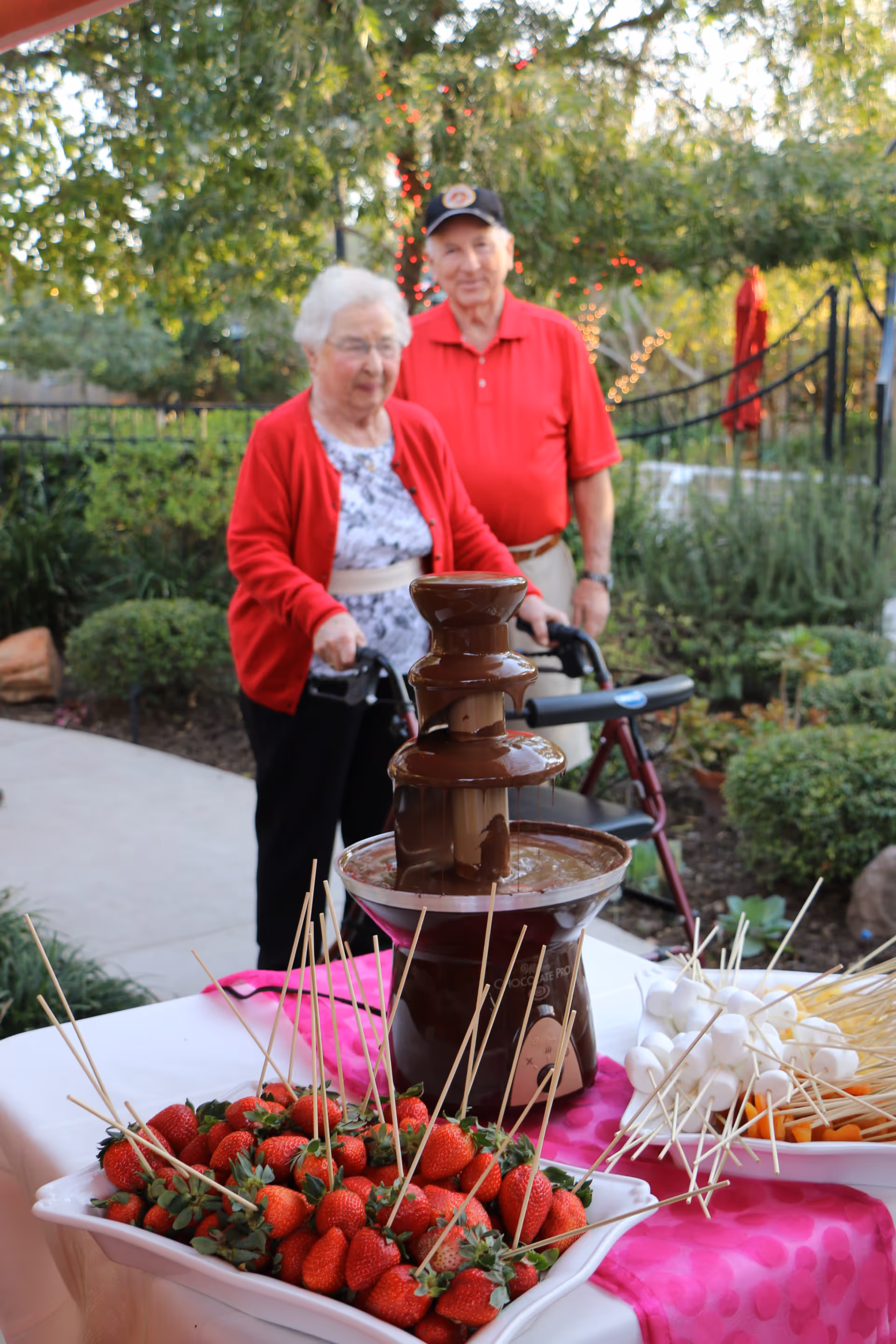 An elderly woman and man standing outdoors near a table with a chocolate fountain, strawberries, marshmallows, and other dipping items. The woman is using a walker and both are dressed in red tops. The background shows greenery and string lights wrapped around a tree.