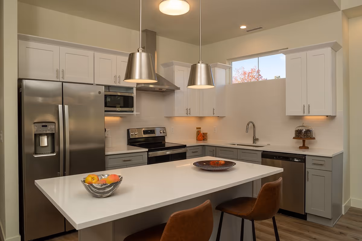 Modern kitchen with white and gray cabinets, stainless steel refrigerator, oven, microwave, and dishwasher. A white island countertop with two brown chairs, a bowl of fruit, and a decorative plate. Two pendant lights hang above the island. A window above the sink shows a view of trees outside.