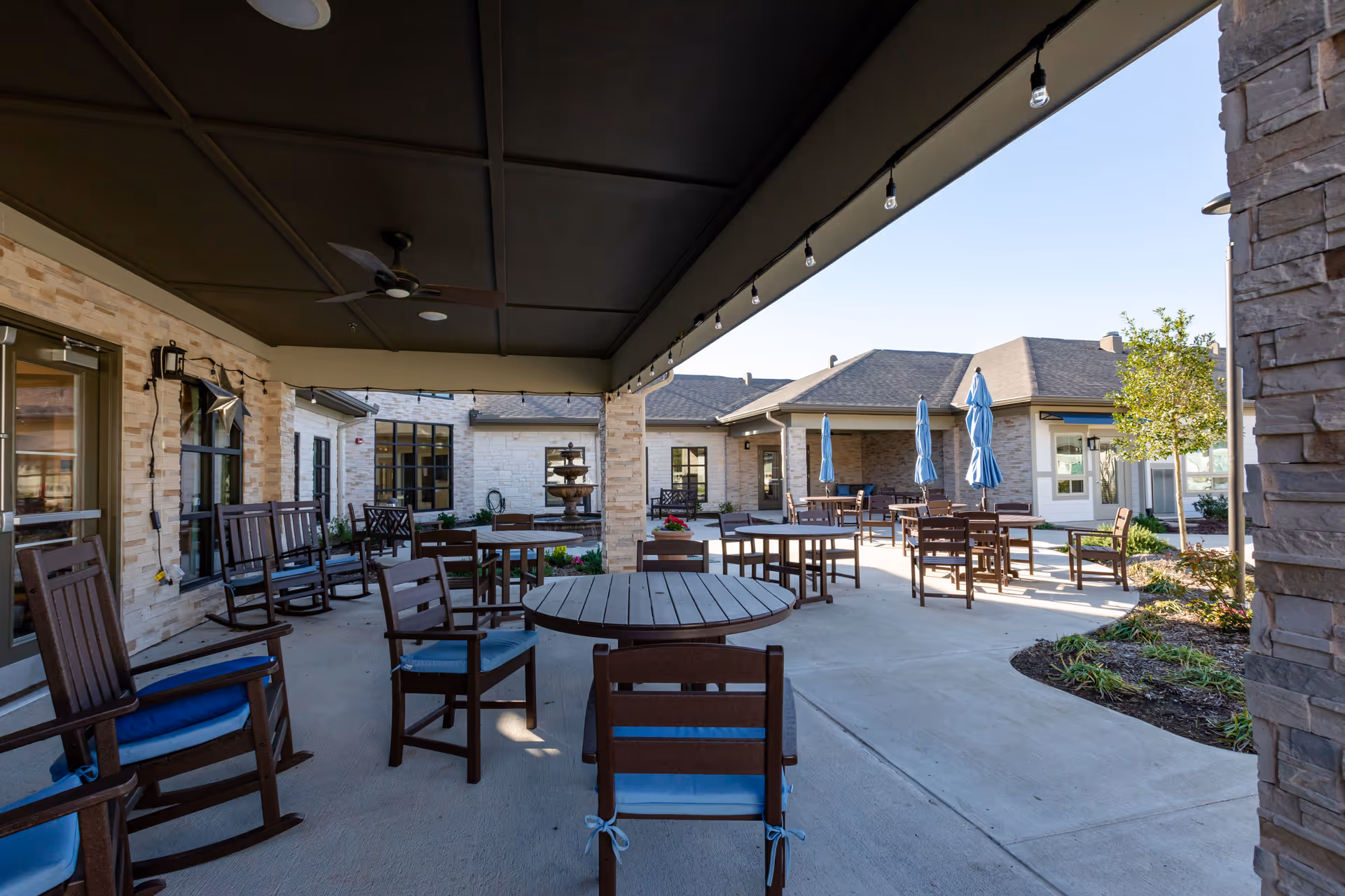 Outdoor covered patio area with multiple wooden tables and chairs, some with blue cushions. There are ceiling fans and string lights hanging from the ceiling. Beyond the covered area, there are more tables with closed blue umbrellas and a stone building with windows and doors. A small garden area with plants and a tree is visible on the right side.