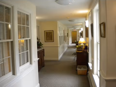 Carpeted interior hallway in a senior living facility lined with windows, framed art, lamps, and console tables.